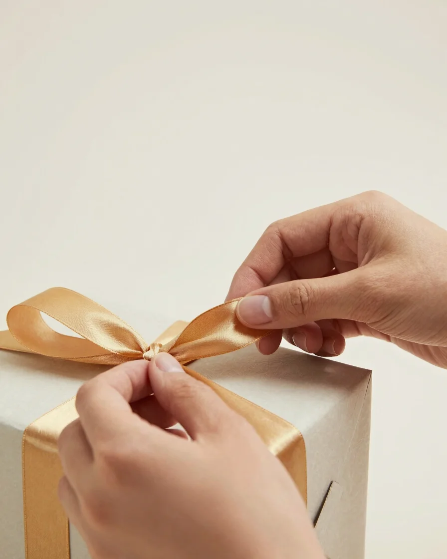 Close-up of hands carefully tying a gold ribbon around a gift box, soft studio lighting, minimalist background, crisp de